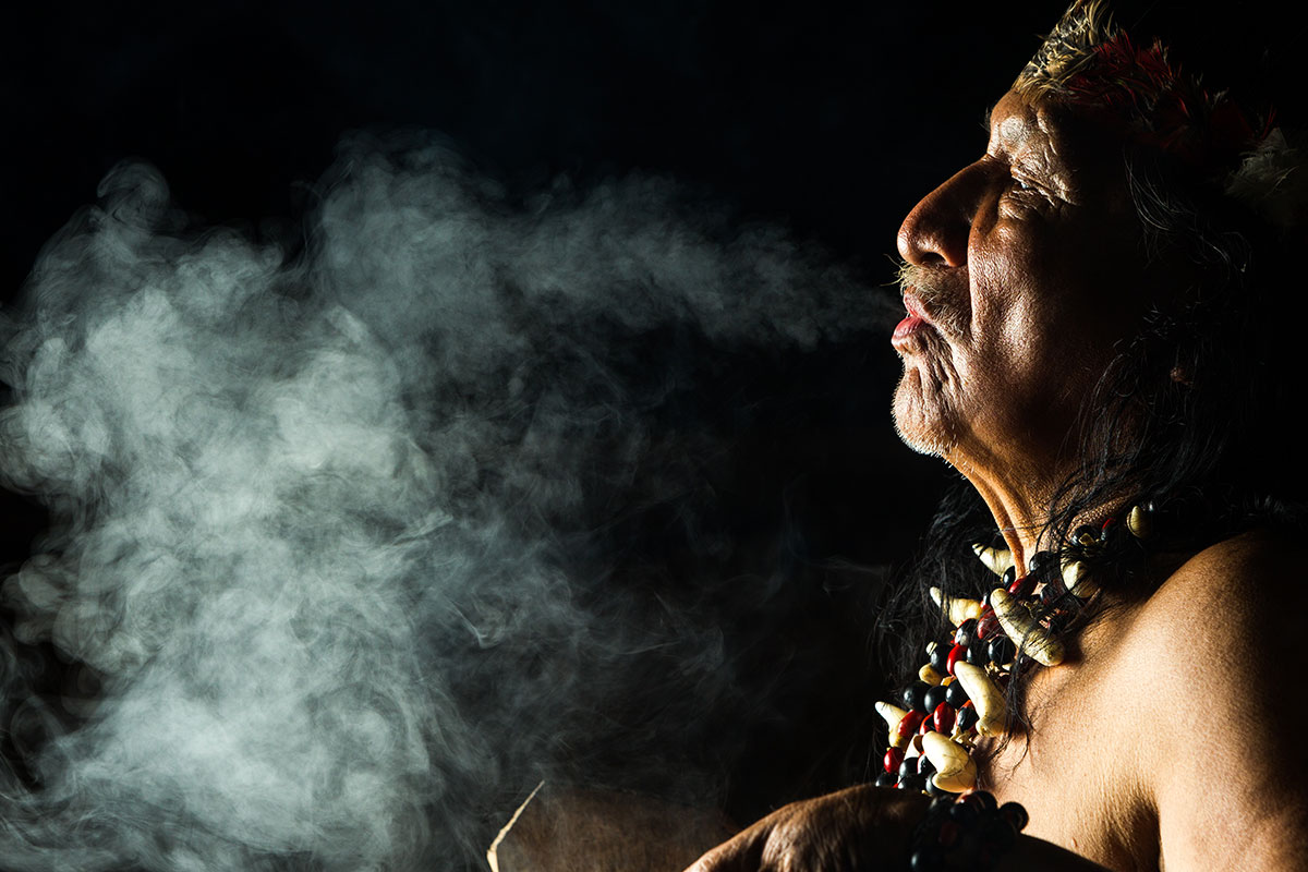an older male wearing jewelry and a headdress and blowing smoke into a black background