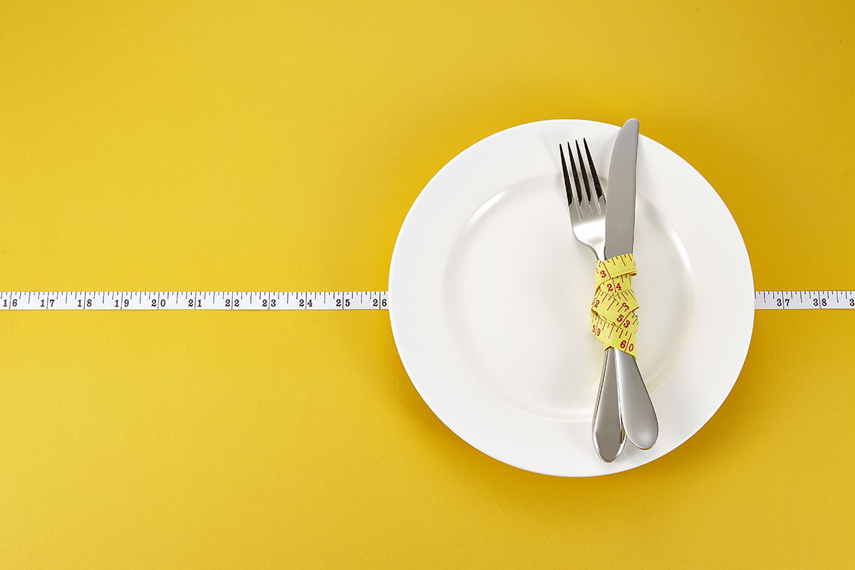 A fork is sitting on a plate, wrapped in a tape measure, set against a yellow background