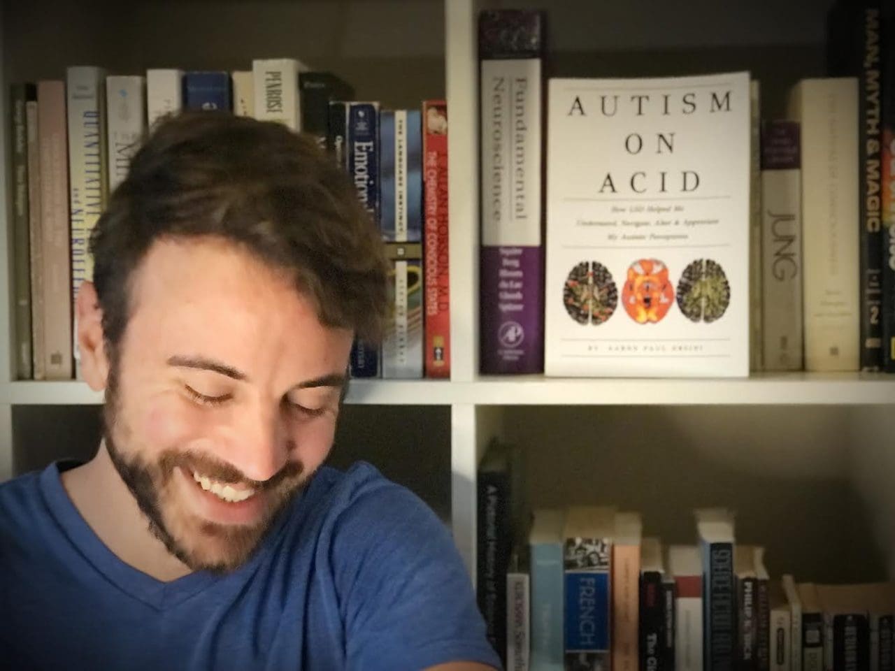 Aaron, a man with brown hair and a beard looks down, smiling, next to a book shelf with his book Autism on Acid displayed