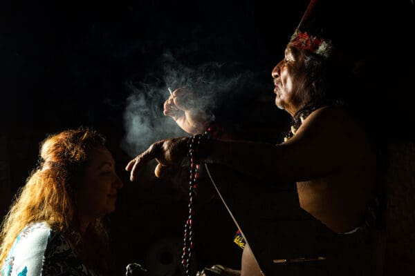 A shaman blows smoke on a white person in a dark room